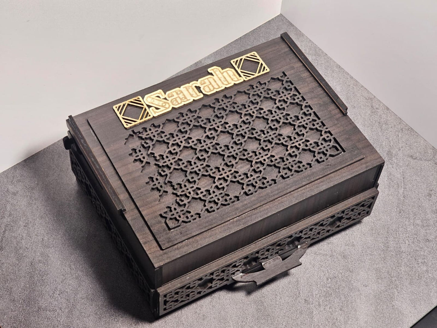 Closed chocolate box made of walnut wood with laser-cut design and custom gold nameplate on top, shown on a grey background.