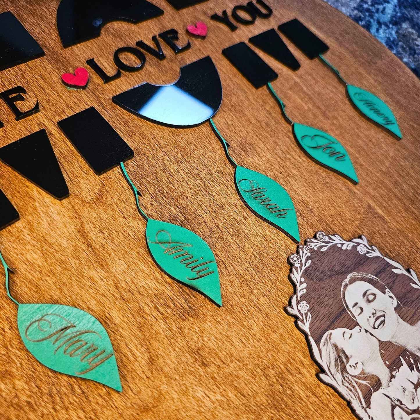 Close-up of black acrylic MOM text and green leaf name tags on brown oak wood – engraved photo below